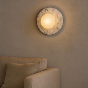 May include: A round wall sconce with a marble-like pattern and a central, glowing orb. The sconce is mounted on a neutral-toned wall, with a beige sofa visible in the foreground. The light fixture provides ambient lighting.