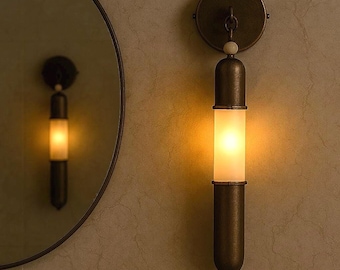 May include: A bronze-colored wall sconce with a cylindrical frosted glass shade, emitting a warm, yellow glow. The sconce has a circular mounting plate and a decorative top element. The sconce is mounted on a textured, light-colored wall.