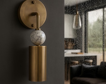 May include: A brass wall sconce with a circular backplate and a cylindrical base. A marble sphere sits between the base and the curved arm. The sconce is mounted on a dark gray wall, with a blurred interior scene in the background.