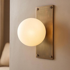 May include: A modern wall sconce featuring a spherical, frosted glass shade emitting a warm glow. The light is mounted on a rectangular, brushed brass backplate, secured with four visible screws. The fixture is designed for indoor use.
