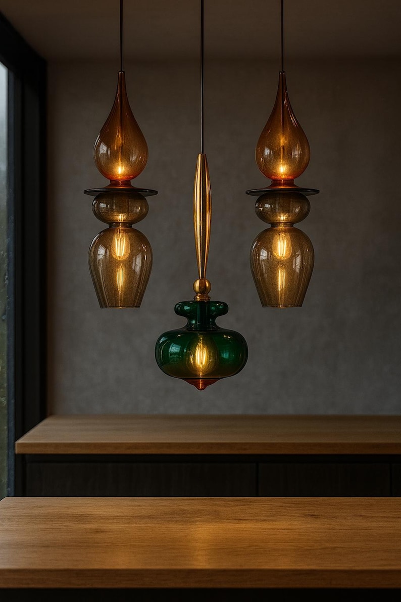 Peut inclure: Trois lampes suspendues avec des abat-jours en verre ambr&eacute; et vert. Les lampes ont une forme de goutte d'eau en haut et un fond bulbeux, avec un accent m&eacute;tallique dor&eacute;. Les lampes sont suspendues au plafond, &eacute;clairant une surface en bois.