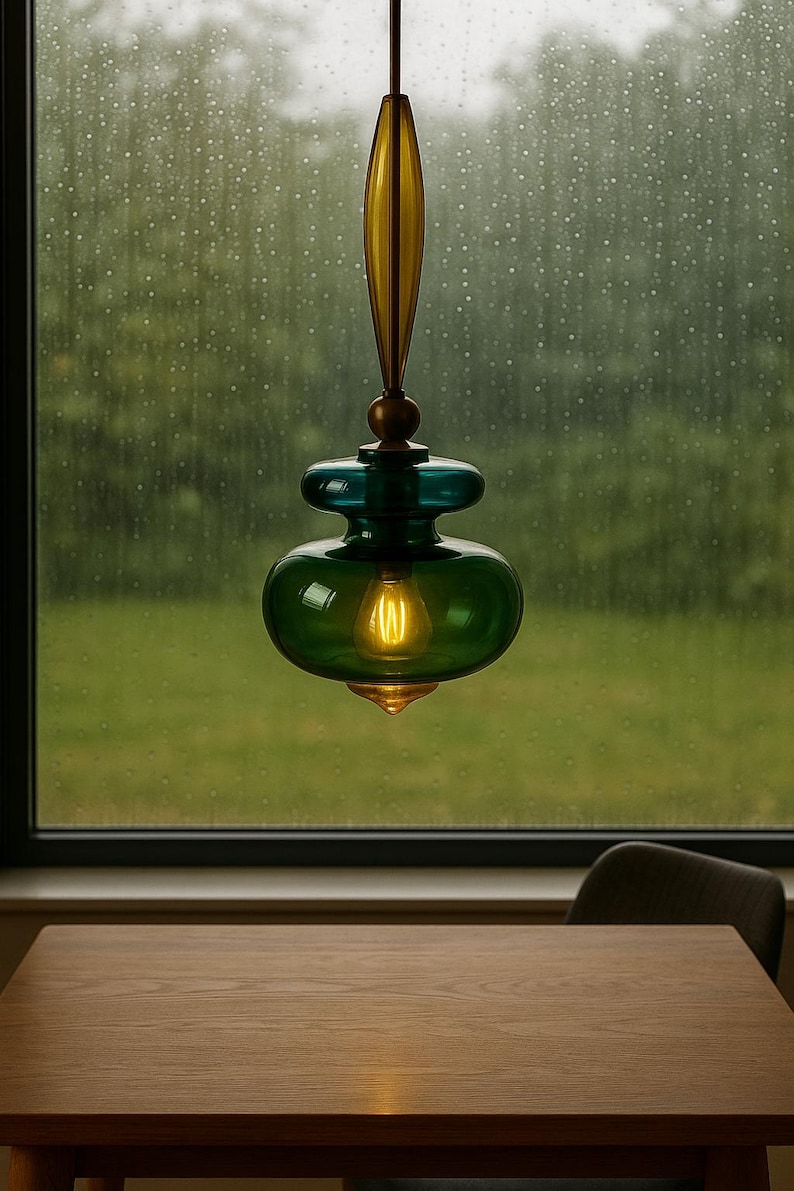 Peut inclure: Un luminaire suspendu avec un abat-jour en verre vert et une ampoule chaude. Le luminaire a des accents sup&eacute;rieurs et inf&eacute;rieurs de couleur laiton. La lumi&egrave;re est suspendue devant une fen&ecirc;tre avec une vue d'arri&egrave;re-plan verte et floue.