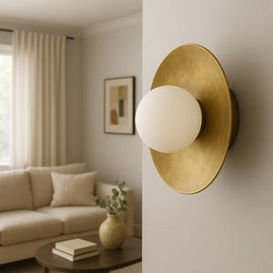 May include: A modern wall sconce with a brass finish and a spherical, frosted glass light. The sconce is mounted on a light gray wall, with a living room setting in the background. The design is minimalist and elegant.