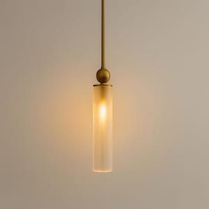 May include: A cylindrical pendant light fixture featuring a frosted glass shade that emits a warm, soft glow. The light is suspended by a slender, matte brass-toned metal rod, which connects to a spherical brass accent piece above the glass cylinder.