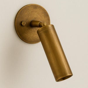 May include: A brass-toned wall-mounted spotlight fixture. The fixture has a circular base with a hexagonal bolt and a cylindrical, adjustable light head. The finish is a warm, metallic gold.
