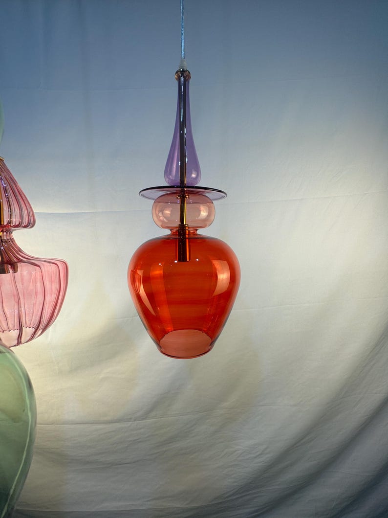 May include: A red, purple, and clear glass pendant light fixture with a unique, modern design. The light fixture has a round, bulbous shape with a pointed top and a small, round base.