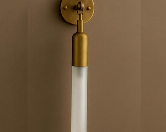 May include: A wall-mounted light fixture with a round, brass-coloured base and a cylindrical frosted glass shade. The fixture has a simple, elegant design, suitable for various interior styles. The overall height is approximately 30 cm.