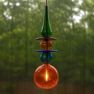 May include: A colorful, handcrafted glass pendant light fixture. The design features stacked, spherical and disc-shaped elements in shades of orange, blue, green, and brown. The light bulb inside the orange glass sphere is illuminated, creating a warm glow.