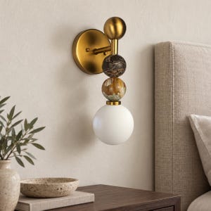 Puede incluir: Un aplique de pared con una placa circular de latón y un brazo vertical. La lámpara incluye una esfera de latón, una esfera de mármol marrón oscuro, una esfera de piedra beige y blanca, y un globo de vidrio blanco.
