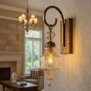 May include: A wall-mounted light fixture featuring a curved brass arm and a rectangular backplate with an intricate pattern. The lamp includes a clear, ribbed glass shade with a teardrop shape and a warm, glowing filament bulb inside.