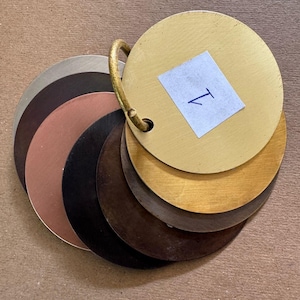 May include: A set of seven round metal samples in various shades of brown, gold, and silver. The samples are attached to a ring and labelled with the letter "T".