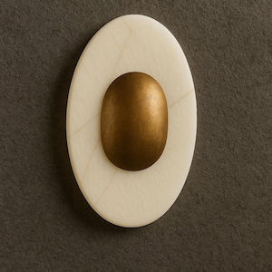 May include: Oval-shaped decorative object with a cream-colored marble base and a central, bronze-colored oval. The marble has subtle veining, and the bronze element has a smooth, metallic finish. The object is set against a dark gray background.