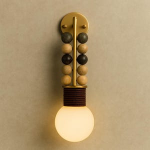 May include: A wall-mounted light fitting featuring a vertical brass rod adorned with two parallel columns of alternating dark grey and light tan spherical beads. Below the beads, a dark brown wrapped cylindrical base supports a round white bulb.