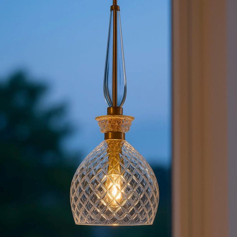 May include: A clear glass pendant light with a diamond pattern shade. The light fixture has a gold-colored metal base and a clear, elongated glass top. The light is illuminated, casting a warm glow.