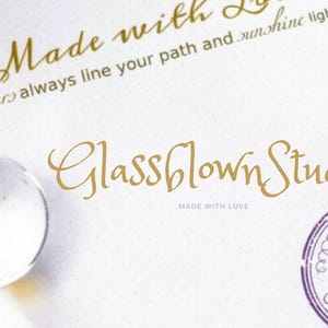 Peut inclure: Une carte blanche avec une &eacute;criture dor&eacute;e qui dit "Made with Love" et "Glassblown Studio". Un globe de verre transparent est dans le coin inf&eacute;rieur gauche. Un tampon violet avec une main tenant un stylo est dans le coin inf&eacute;rieur droit.