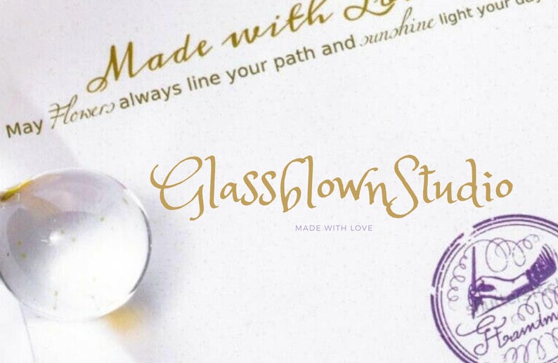 Peut inclure: Une carte blanche avec une &eacute;criture dor&eacute;e qui dit "Made with Love" et "Glassblown Studio". Un globe de verre transparent est dans le coin inf&eacute;rieur gauche. Un tampon violet avec une main tenant un stylo est dans le coin inf&eacute;rieur droit.