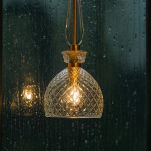 May include: A pendant light fixture with a clear, diamond-patterned glass shade and a gold-colored metal frame. The light is illuminated, casting a warm glow. The background is a dark, blurred view of a window with raindrops.