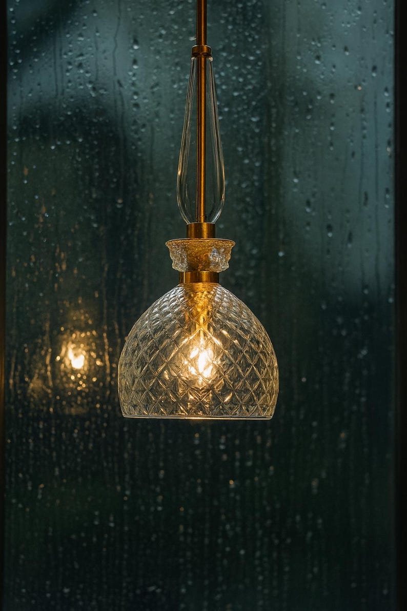 May include: A pendant light fixture with a clear, diamond-patterned glass shade and a gold-colored metal frame. The light is illuminated, casting a warm glow. The background is a dark, blurred view of a window with raindrops.