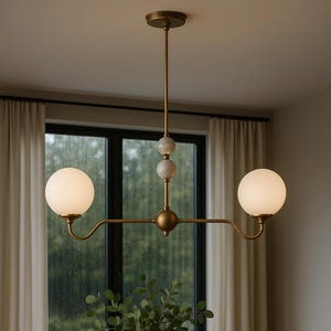Puede incluir: Una lámpara de araña moderna de latón con dos pantallas esféricas de vidrio esmerilado. La lámpara tiene una varilla vertical central con detalles de mármol y un brazo horizontal que se extiende a cada luz. El diseño es minimalista y elegante.