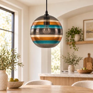 Puede incluir: Una lámpara colgante esférica de cristal con bandas horizontales de colores ámbar, turquesa y gris translúcidos. El globo está suspendido por un cable negro fino y contiene una bombilla transparente visible a través del cristal.