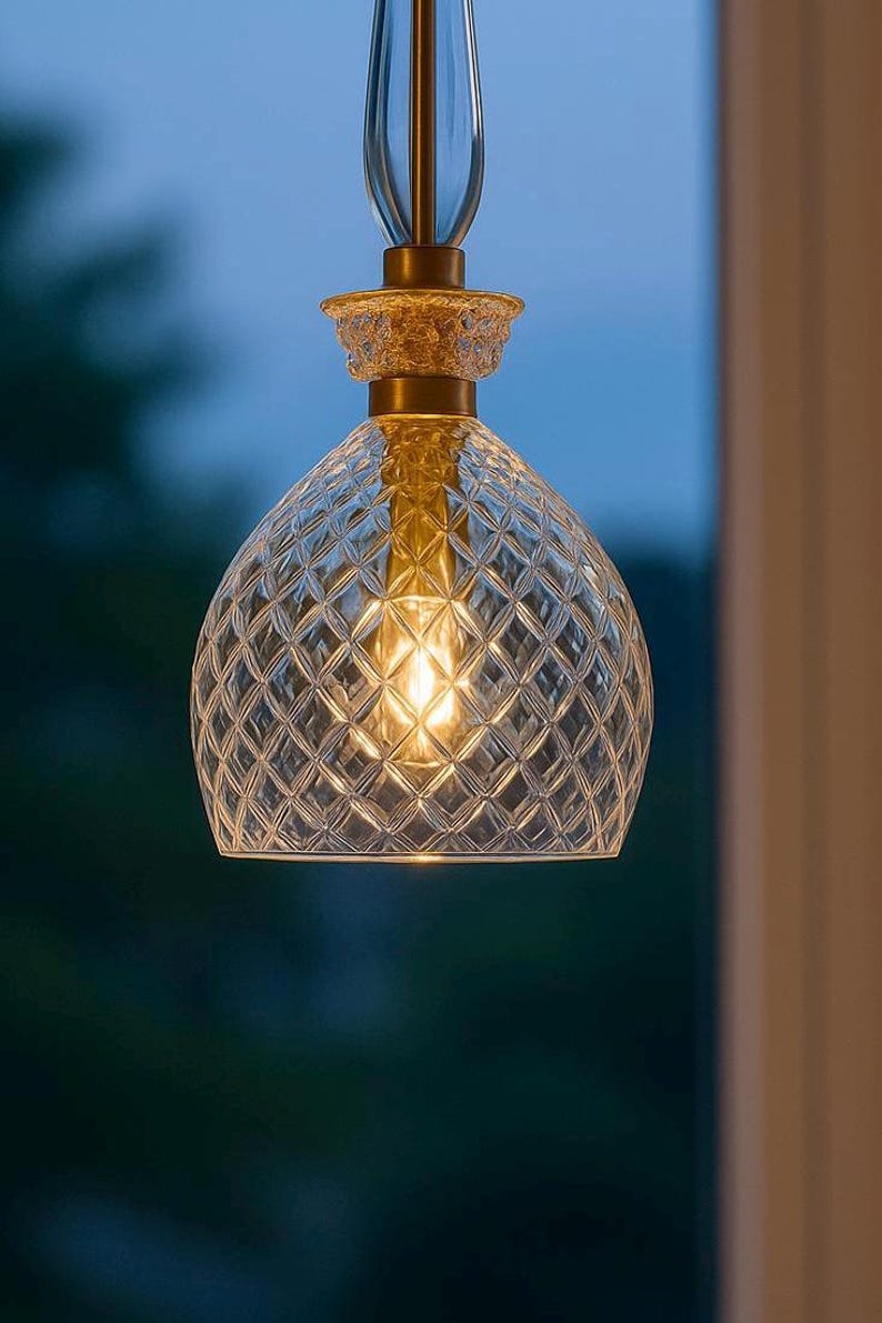 May include: A clear glass pendant light with a diamond pattern shade. The light fixture has a gold-colored metal base and a clear glass stem. The light is illuminated, casting a warm glow. The background is a blurred outdoor scene.
