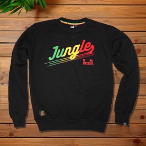 Peut inclure: Un sweat-shirt noir à manches longues avec le texte "Jungles IS MASSIVE." en dégradé vert, jaune et rouge. Le motif comprend des lignes horizontales sous le mot "Jungles" et une petite icône de lion à côté du mot "MASSIVE."
