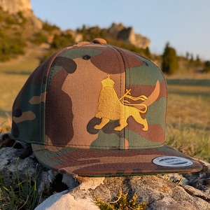 May include: Camouflage-patterned baseball cap with a flat brim. The cap features an embroidered gold lion design on the front. The hat is resting on a rock, with a natural outdoor background.