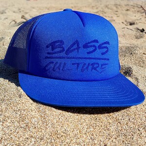 Op de afbeelding: Een koninklijk blauwe truckercap met een zwart mesh achterpaneel. De pet heeft een zwart geborduurd logo met de tekst "BASS CULTURE" in een gestileerd lettertype.
