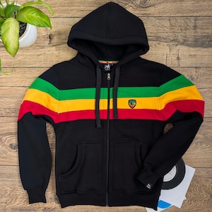 Op de afbeelding: Een zwarte hoodie met ritssluiting en een horizontaal streeppatroon in groen, geel en rood over de borst en mouwen. Het kledingstuk heeft een capuchon met trekkoord, twee voorzakken en een klein schildvormig embleem op de linkerborst.