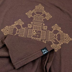May include: A brown t-shirt with a gold geometric design featuring a lion. The shirt has a tag that reads "Roots & Culture".