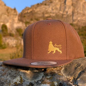 May include: A brown snapback hat with a flat brim. The hat features a gold embroidered lion emblem on the front. The hat is sitting on a stone surface with a blurred background of a mountain.