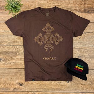 May include: A brown t-shirt with a gold geometric cross design and the text "አትሐበለቅ" printed on the front. A black baseball cap with the text "ROOTS REALITY CULTURE" is also shown.
