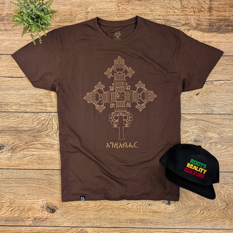May include: A brown t-shirt with a gold geometric cross design and the text "አትሐበለቅ" printed on the front. A black baseball cap with the text "ROOTS REALITY CULTURE" is also shown.