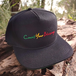 May include: Black baseball cap with a green, yellow, and red embroidered design that says "Count Your Blessings".