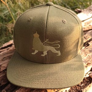 May include: Olive green baseball cap with a flat brim. The cap features an embroidered lion design in tan and gold on the front. The cap has a structured crown and a curved brim.