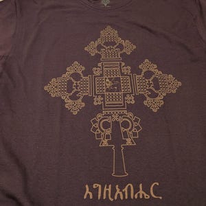 May include: A dark brown t-shirt with a gold-colored Ethiopian Orthodox cross design. The cross is intricate and detailed, with a small figure in the center. The text "እግዚአብሔር" is printed below the cross.