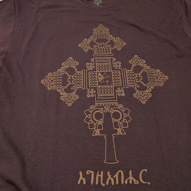 May include: A dark brown t-shirt with a gold-colored Ethiopian Orthodox cross design. The cross is intricate and detailed, with a small figure in the center. The text "እግዚአብሔር" is printed below the cross.