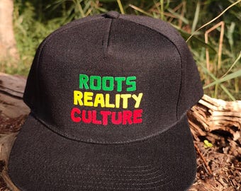 May include: Black snapback hat with the words "ROOTS REALITY CULTURE" embroidered in green, yellow, and red. The hat has a flat brim and a structured crown. The hat is sitting on a piece of wood.