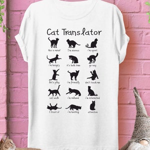 Puede incluir: Camiseta blanca con un gráfico de traductor de gatos negro. El gráfico muestra diferentes poses de gatos y sus significados correspondientes, como "¡esto es mío!" y "estoy cazando".