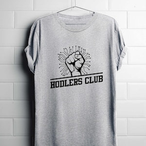 Può includere: Una maglietta grigia con una grafica nera di un pugno all'interno di un design di circuito stampato. Il testo "HODLERS CLUB" è stampato sotto la grafica. La maglietta è appesa a un appendiabiti contro un muro piastrellato bianco.