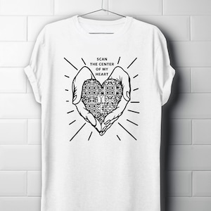 Può includere: Maglietta bianca con un design grafico in bianco e nero di due mani che tengono un cuore con un codice QR al suo interno. Il testo "SCAN THE CENTER OF MY HEART" è sopra la grafica.