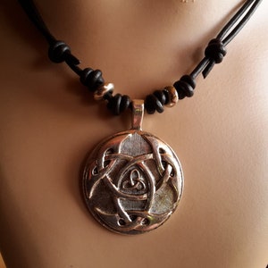 Könnte beinhalten: Halskette mit silberfarbenem Anhänger und keltischem Knotenmuster. Der Anhänger hängt an einer schwarzen Lederschnur mit goldfarbenen Perlen. Die Halskette wird auf einer Schaufensterpuppe präsentiert.