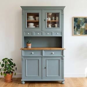 Könnte beinhalten: Ein hoher, schieferblauer Küchenschrank mit einer Arbeitsplatte aus Naturholz, zwei verglasten Obertüren, vier kleinen Schubladen, zwei unteren Schubladen und zwei massiven Basistüren mit weißen Knöpfen, gefüllt mit Keramikgeschirr.