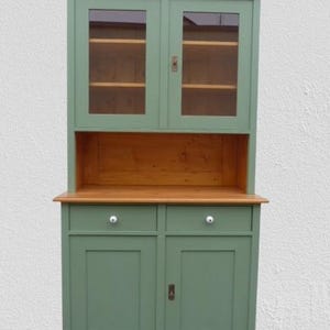 Könnte beinhalten: Ein antiker Schrank in grüner Farbe mit Glastüren und zwei unteren Türen. Der Schrank hat eine Holzplatte und ein Regal im oberen Teil.