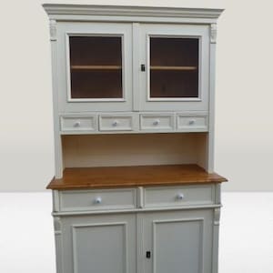 Könnte beinhalten: Eine cremefarbene Holzhutch mit oberen Schränken mit Glasfront und unteren Schranktüren. Sie verfügt über Schubladen mit weißen Knöpfen und eine Arbeitsplatte aus Naturholz. Die Hütte hat dekorative Leisten und ist eine klassische Aufbewahrungslösung.