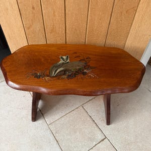 May include: A small wooden stool with a scalloped edge tabletop featuring a painted illustration of a badger surrounded by dark berries and foliage. The stool has a warm brown wood grain finish and is supported by four sturdy, angled wooden legs.