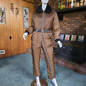 May include: A brown quilted jumpsuit with a zipper down the front and a black fur collar. The jumpsuit has a belt with a silver buckle.