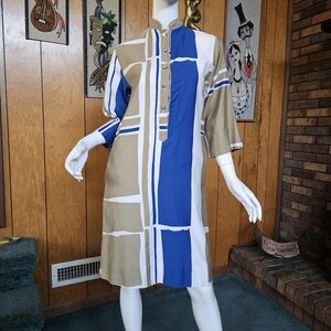 May include: A vintage knee-length dress with a geometric pattern in beige, white, and blue. The dress features a mandarin collar, short sleeves, and a button-down front. The dress is displayed on a mannequin.
