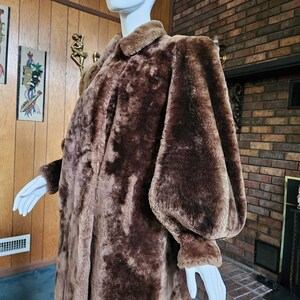May include: A long, brown faux fur coat with a wide collar and large, puffy sleeves. The coat is displayed on a mannequin, showcasing its full length and texture. The coat's color is a rich, warm brown, suggesting a vintage or retro style.