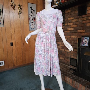 May include: A vintage floral print dress on a mannequin. The dress features a white background with pink roses and green leaves. It has short sleeves, a belted waist, and a knee-length skirt. The dress is displayed on a mannequin with a white torso and arms.